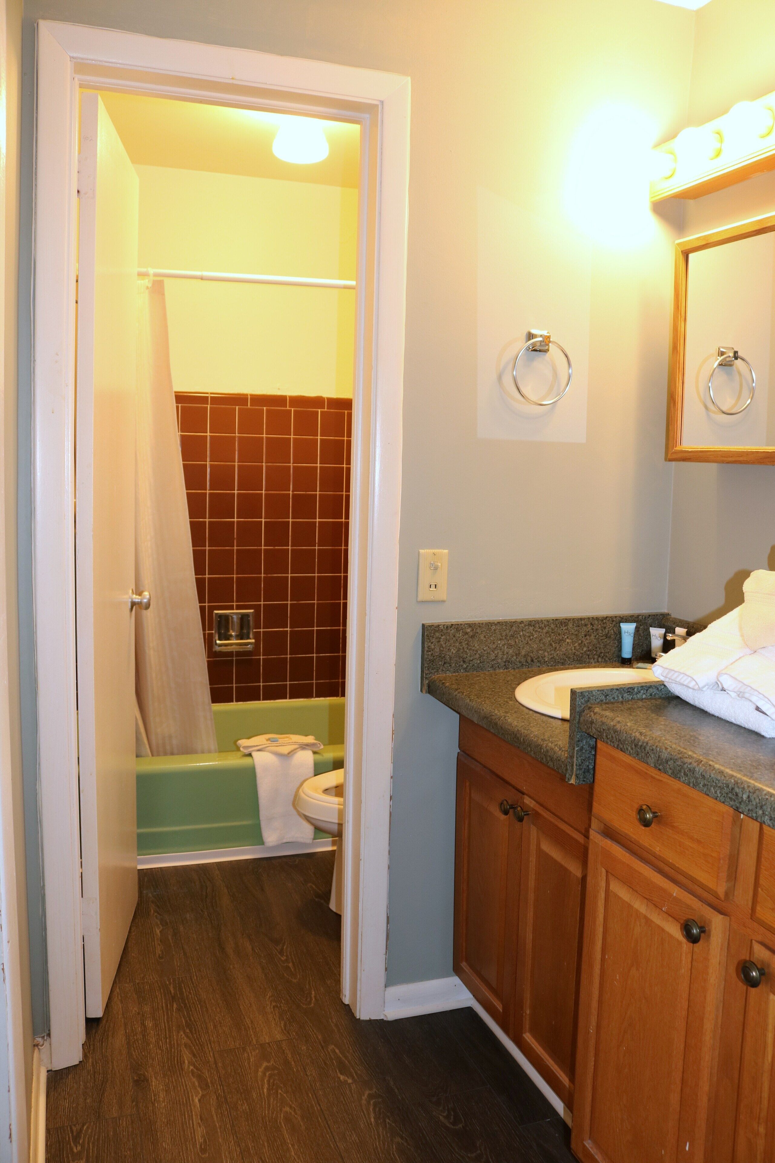 A bathroom with a green bathtub, a white toilet, wooden cabinets, and a mirror with a light above it.