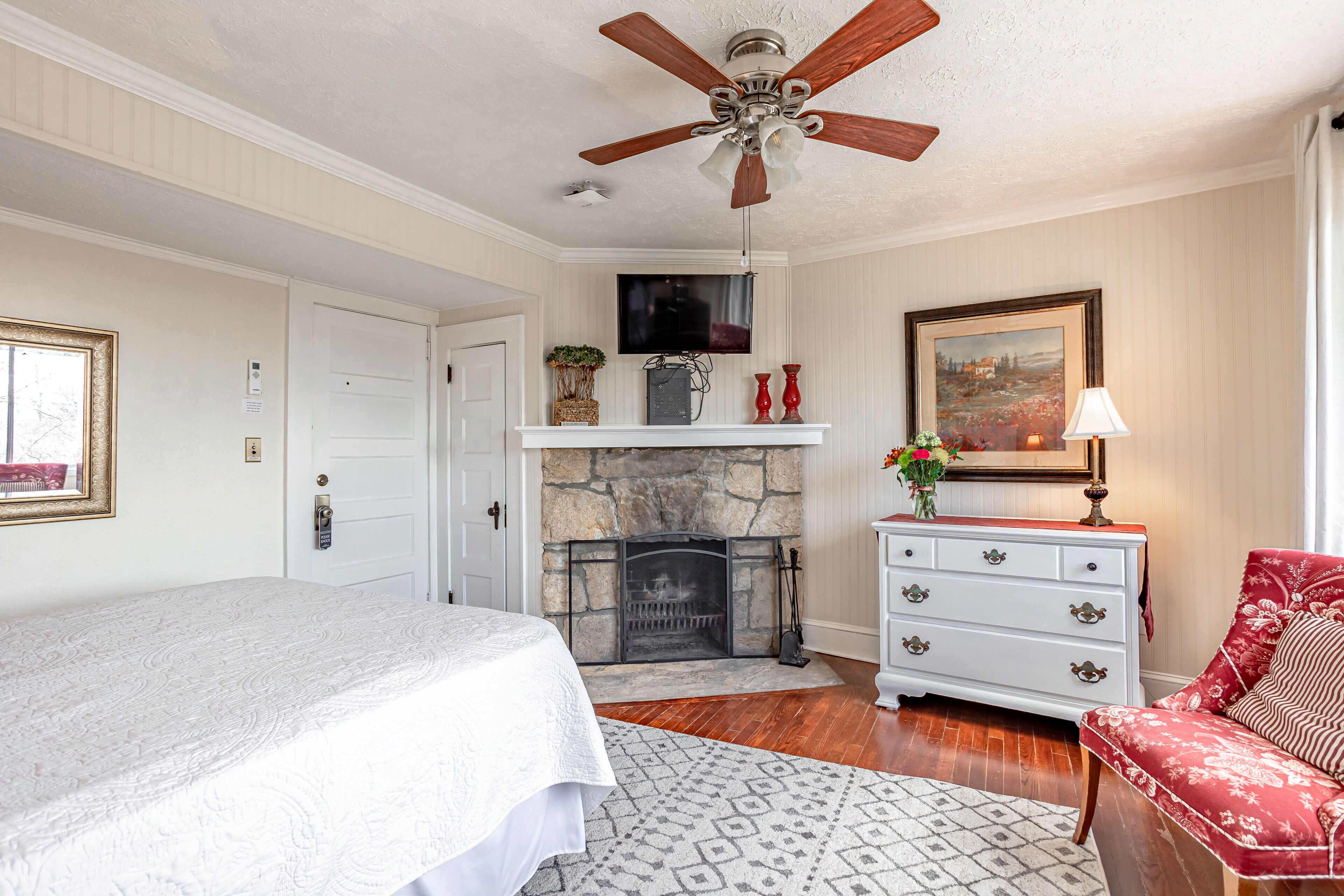 A bedroom with a bed, a fireplace, a television, a ceiling fan, and a red chair.