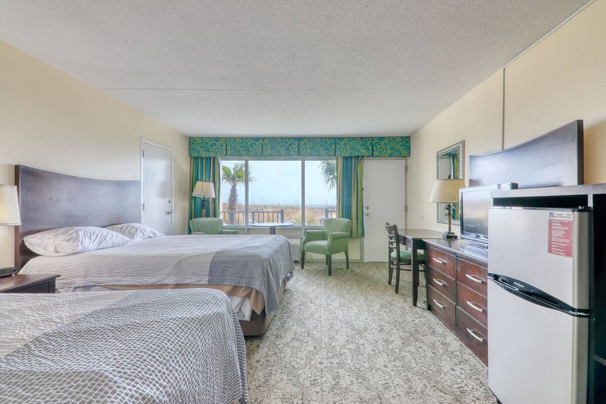 A hotel room with two beds, a desk, a refrigerator, and a view of the beach.