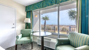 Una habitación con sillas tapizadas a rayas verdes y blancas, una mesita redonda y un ventanal con vistas a la playa y palmeras.