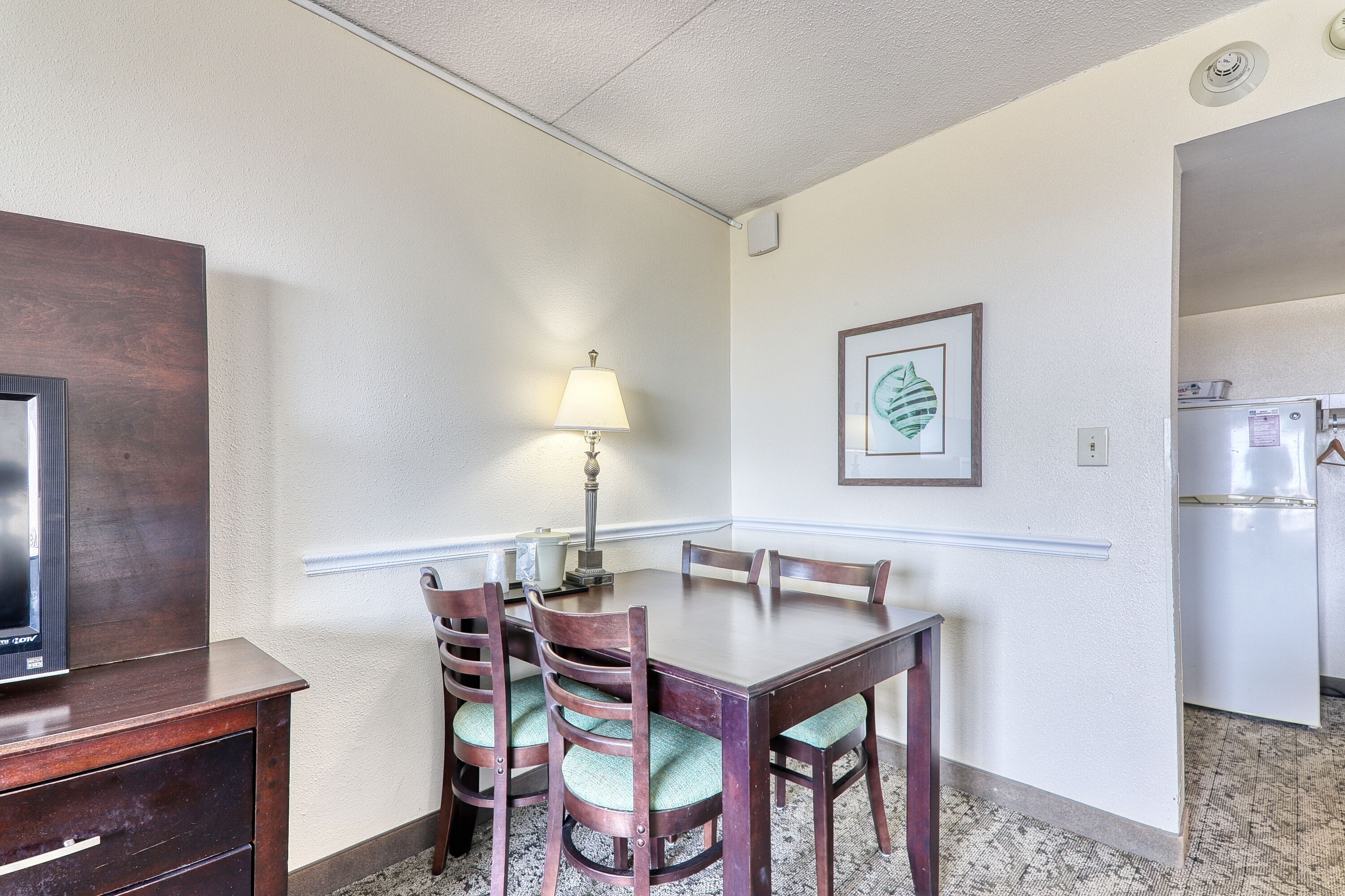 A dining area with a wooden table, four chairs, a lamp, and a framed artwork on the wall.