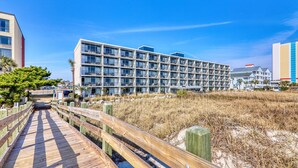 Un hotel moderno con varios pisos y balcones, ubicado cerca de una playa con un paseo marítimo de madera que la conecta.