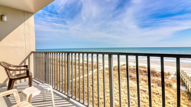 Un balcón con vista a la playa, que incluye una silla de madera, un taburete blanco pequeño y una barandilla de metal.