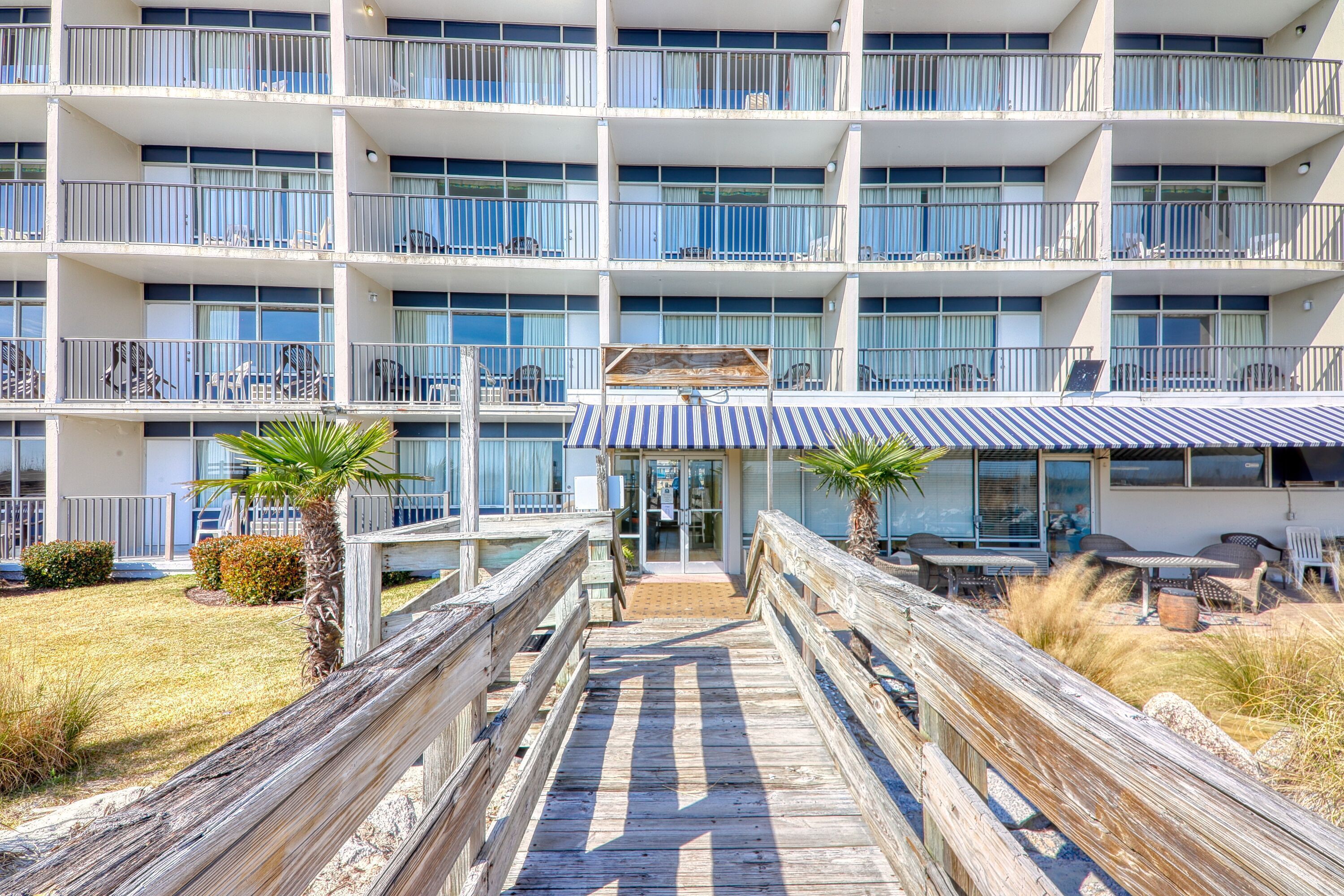 A multi-story hotel with balconies, a covered outdoor seating area, and a wooden boardwalk leading to the entrance.