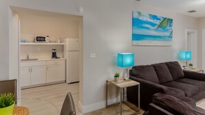 A modern living room with a dark brown sofa, a coffee table, and a blue lamp. There is a kitchen area with a microwave and a coffee machine. A painting of a beach scene is on the wall. - Drift Hotel (Key Largo)