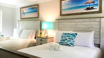 A hotel room with two beds, each with white linens and pillows, a nightstand with a lamp, and artwork depicting beach scenes.