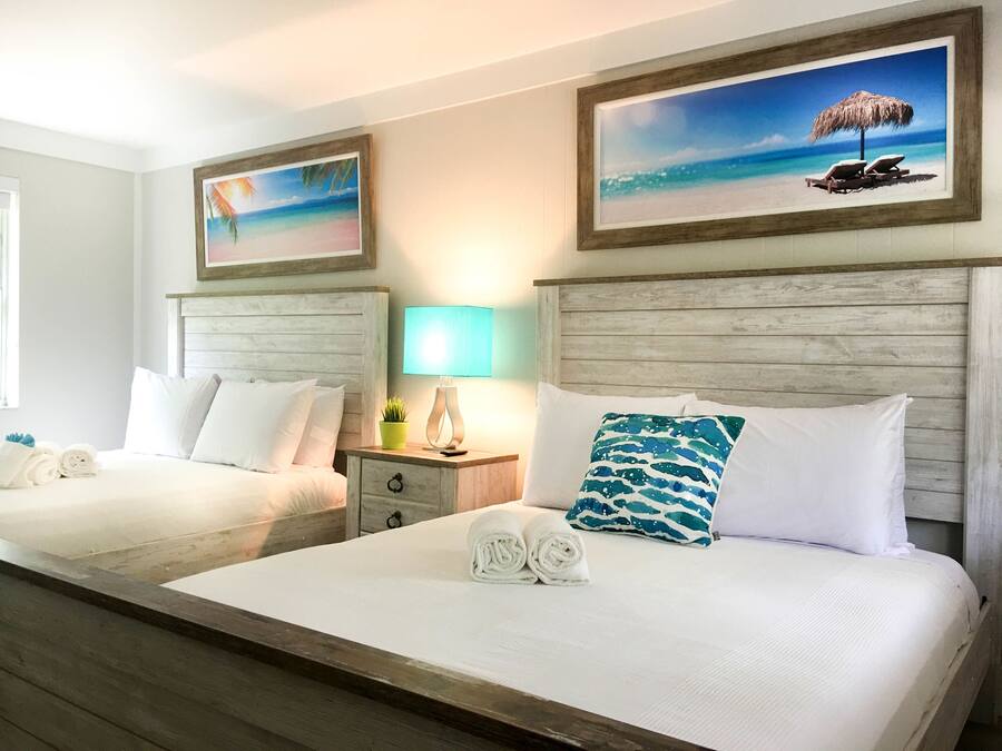A hotel room with two beds, each with white linens and pillows, a nightstand with a lamp, and artwork depicting beach scenes.