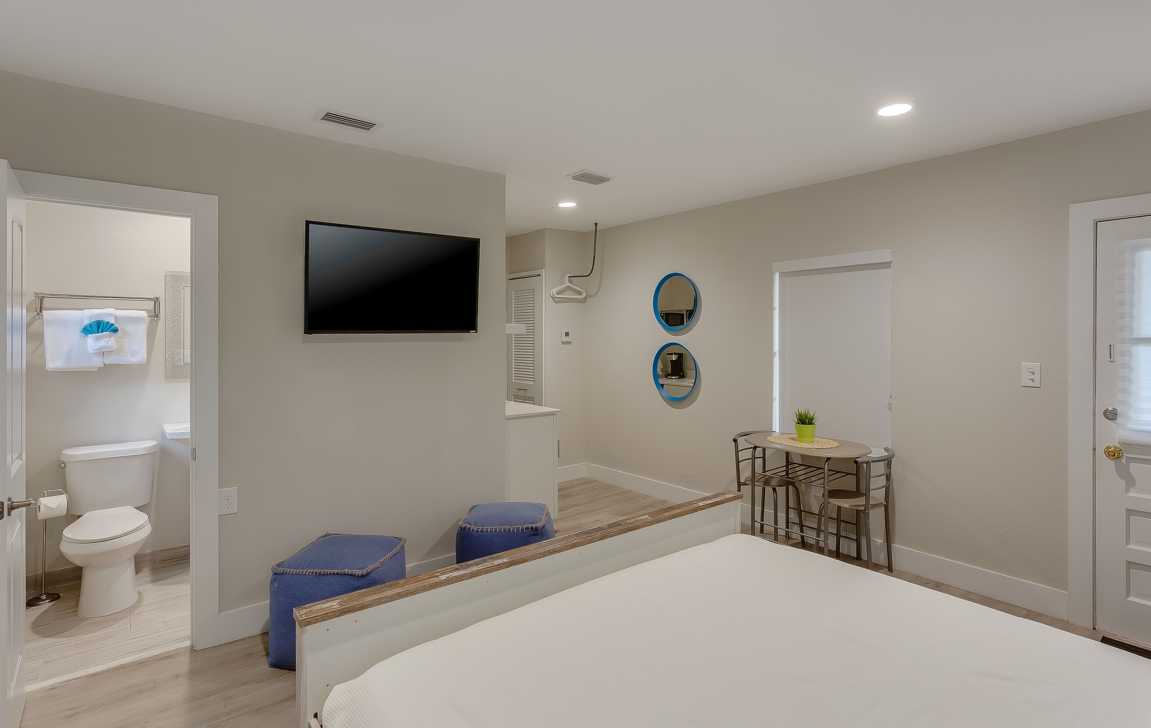 Un dormitorio con cama, televisor, una mesita con sillas y baño con inodoro y perchero para toallas.