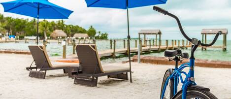 Une plage avec des chaises longues et des parasols bleus, un vélo et une jetée en bois qui s’avance dans l’eau.