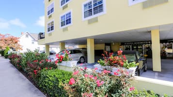Un hotel con fachada amarilla, varias ventanas y un estacionamiento con coches y macizos de flores.