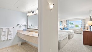 Un baño de hotel con lavamanos, espejo y toallas. Hay un dormitorio con cama y escritorio.