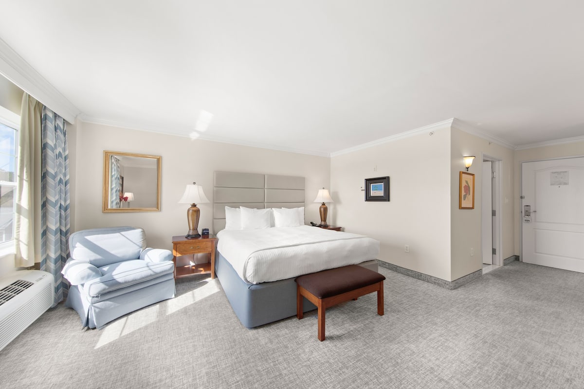 A hotel room with a large bed, a bedside table with lamps, a chair, a bench, and a mirror on the wall.
