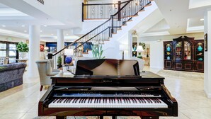 Un piano de cola imponente en un vestíbulo amplio y luminoso, con techo alto, una escalera y una gran estantería.