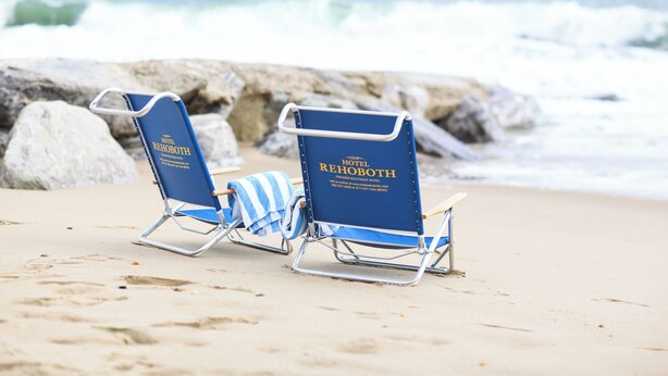 Dos sillones azules con el logotipo de 'Hotel Rehoboth' sobre una playa arenosa con afloramientos rocosos al fondo.