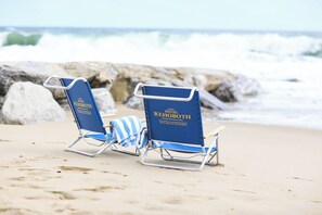 Two blue lounge chairs with "Hotel Rehoboth" branding on a sandy beach with rocky outcrops in the background. - Hotel Rehoboth (Rehoboth Beach)