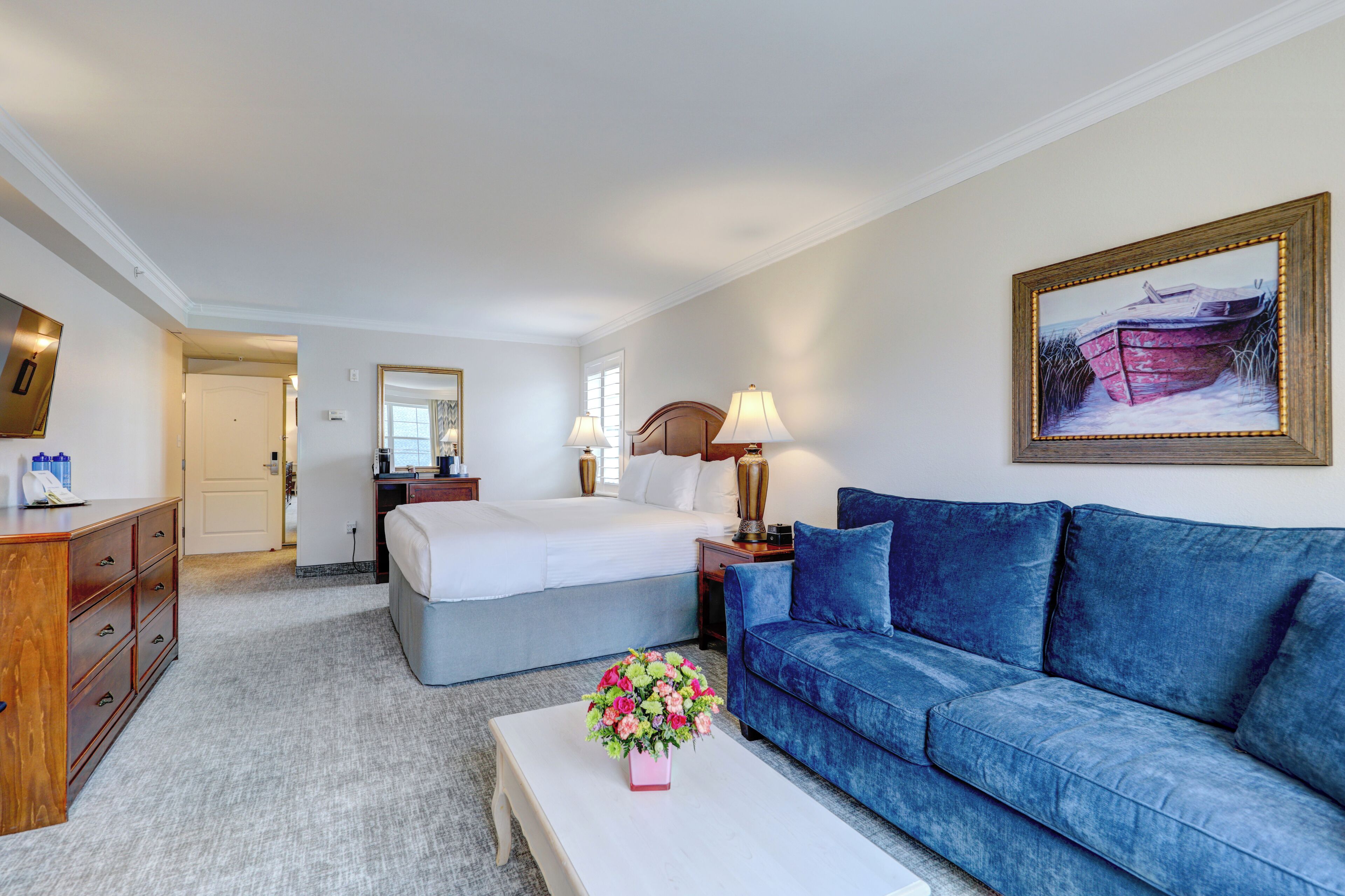 A hotel room with a bed, a blue sofa, a white coffee table with flowers, a wooden dresser, and a painting on the wall.