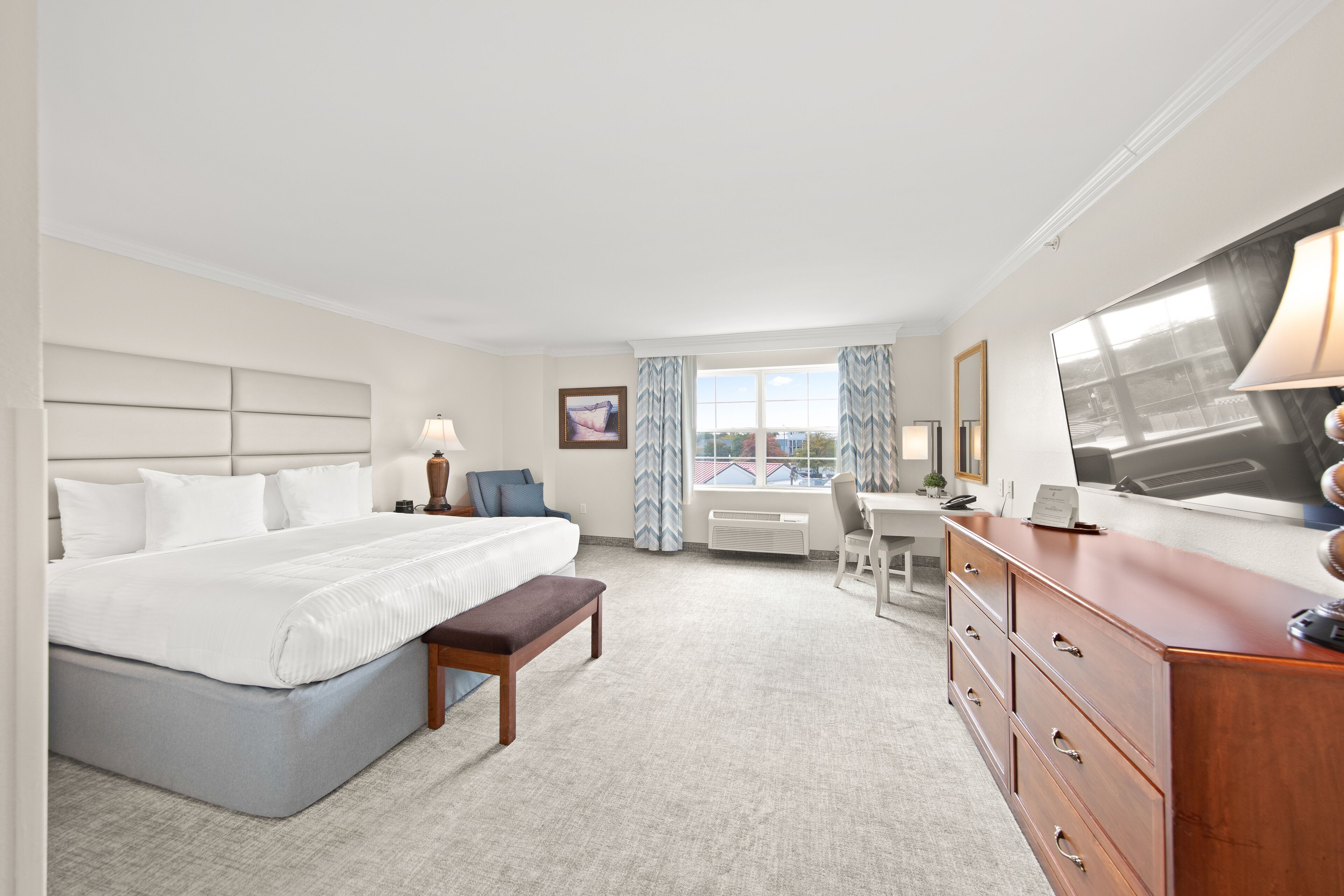 A hotel room with a bed, a desk, a dresser, a bench, a TV, and a window with curtains.