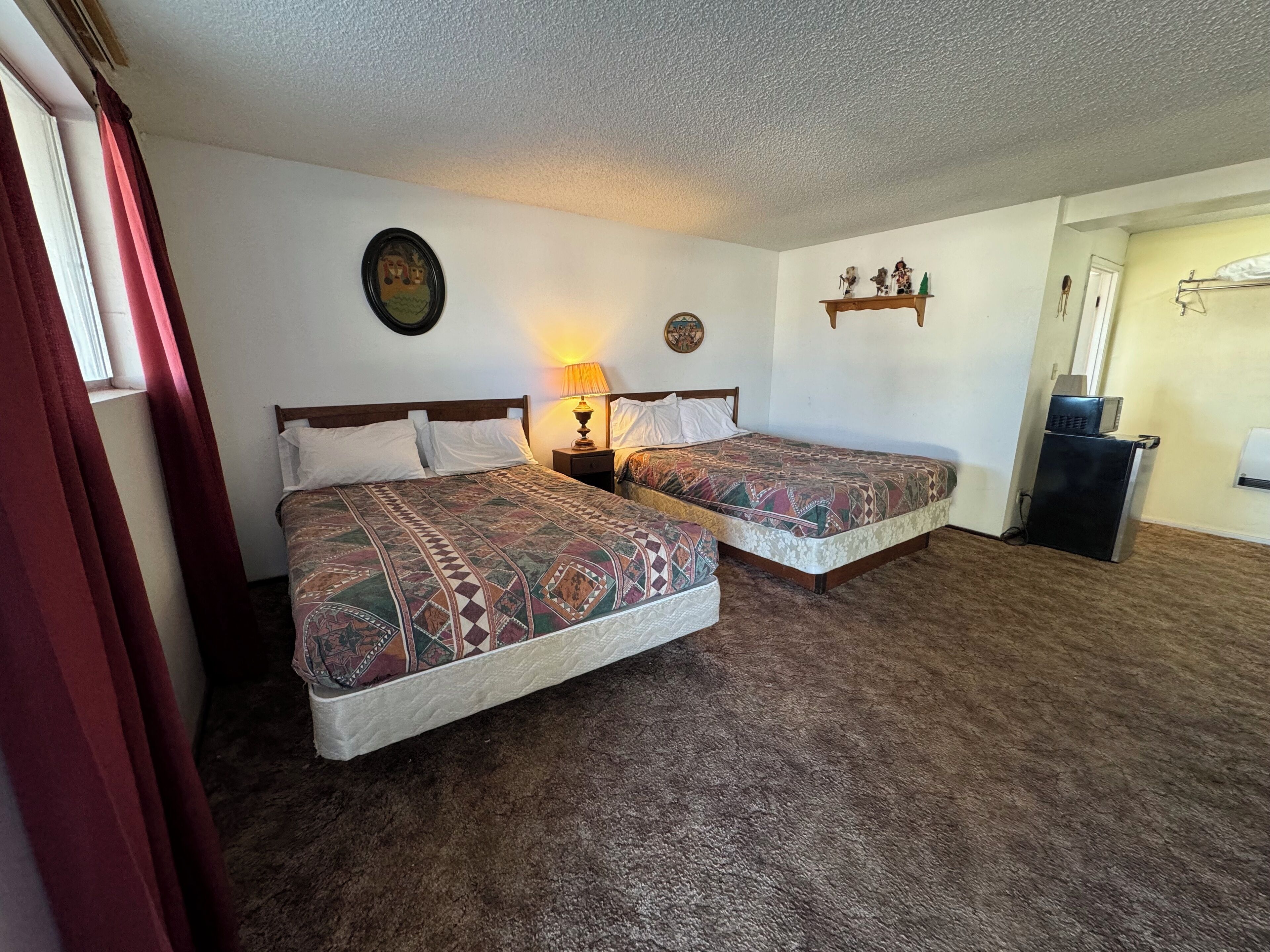 A room with two beds, a nightstand with a lamp, a wall clock, and a small shelf with decorative items.