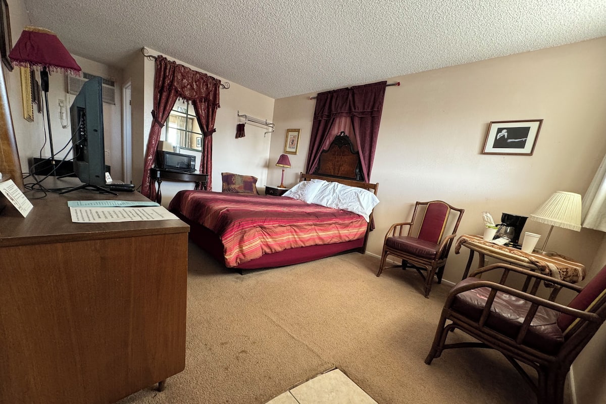 A hotel room with a bed, desk, chair, and a small table with a lamp.