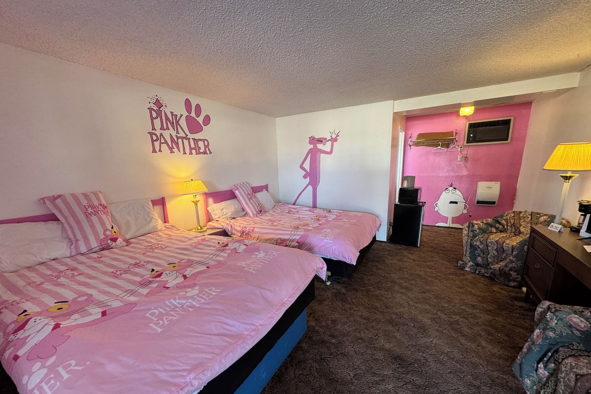 A bedroom with two beds, each with pink bedding and cartoon character prints. There is a pink wall with a 'Pink Panther' sign and a cartoon character cutout. A lamp and a small table are visible.