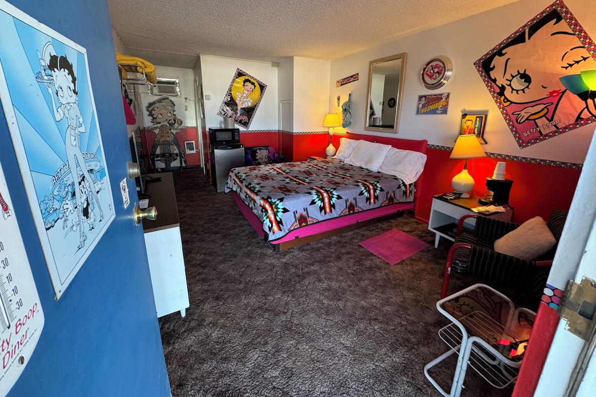 A hotel room with a bed, a desk, a chair, a microwave, a refrigerator, and a wall with various posters and artwork.