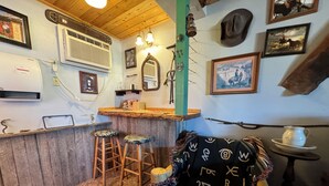 Un rústico espacio de bar con taburetes de madera, una barra y varios objetos decorativos, como un espejo, cuadros enmarcados y un sombrero colgante.