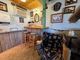 A rustic bar area with wooden stools, a bar counter, and various decorative items including a mirror, framed pictures, and a hanging hat.