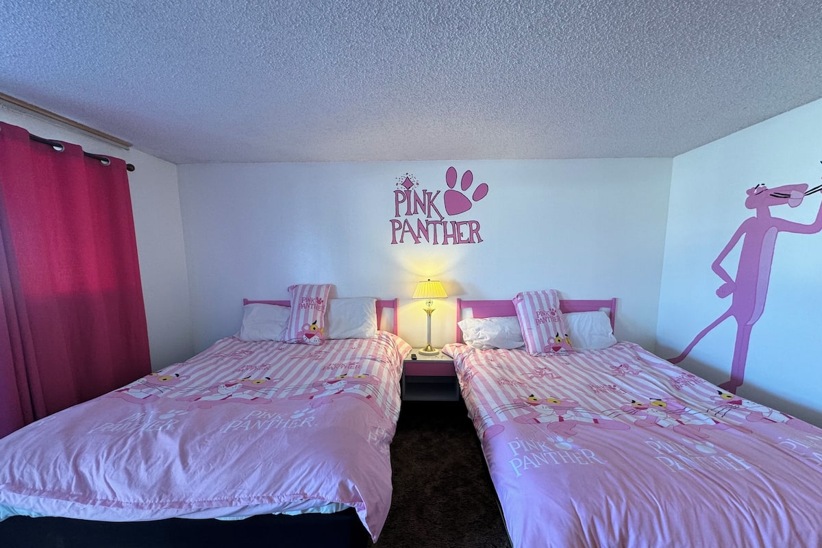 A bedroom with two beds, each with pink bedding and a 'Pink Panther' theme. There is a wall decoration of a pink panther and a silhouette of a person.