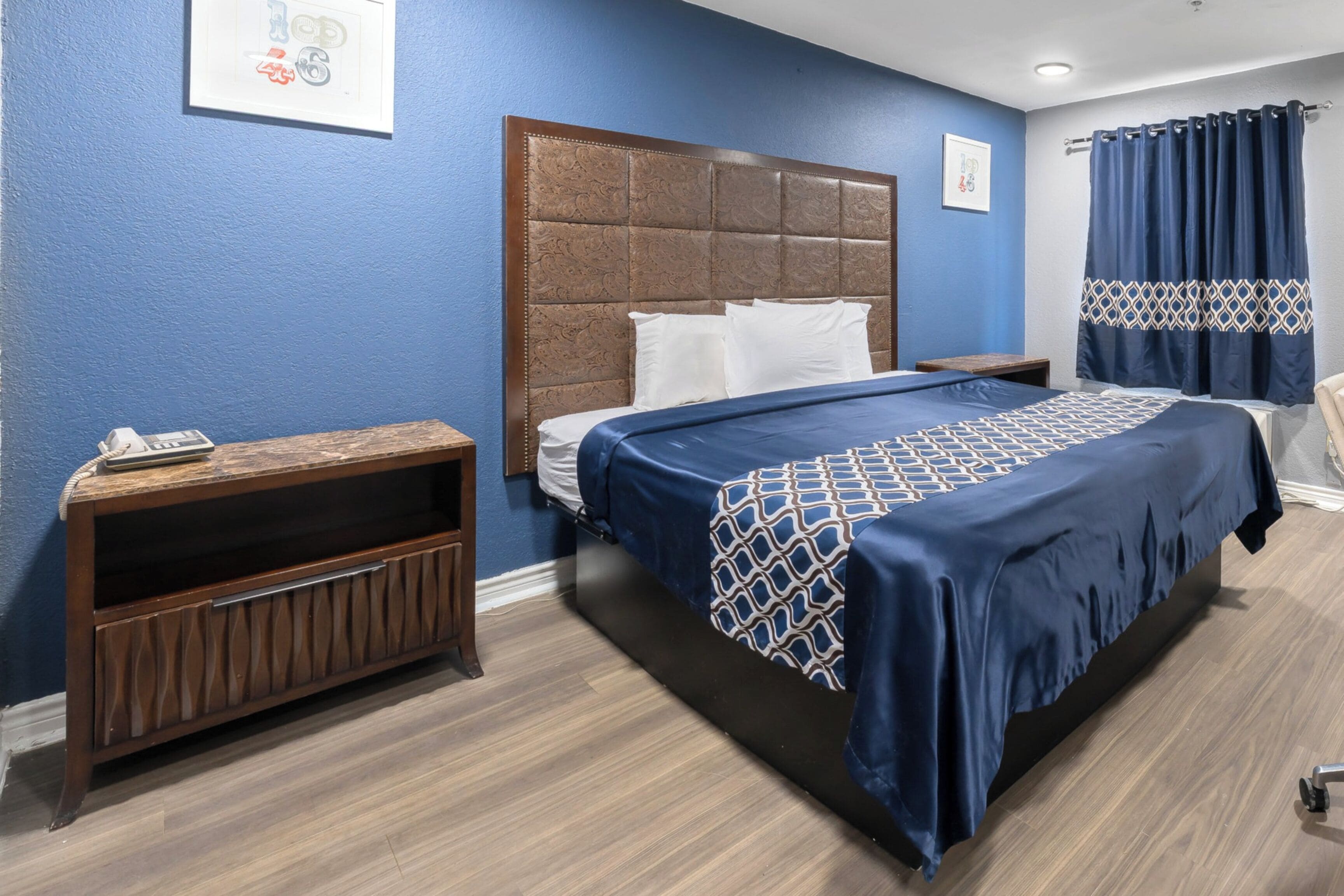 A hotel room with a large bed, a wooden nightstand, a blue wall, and a curtain.