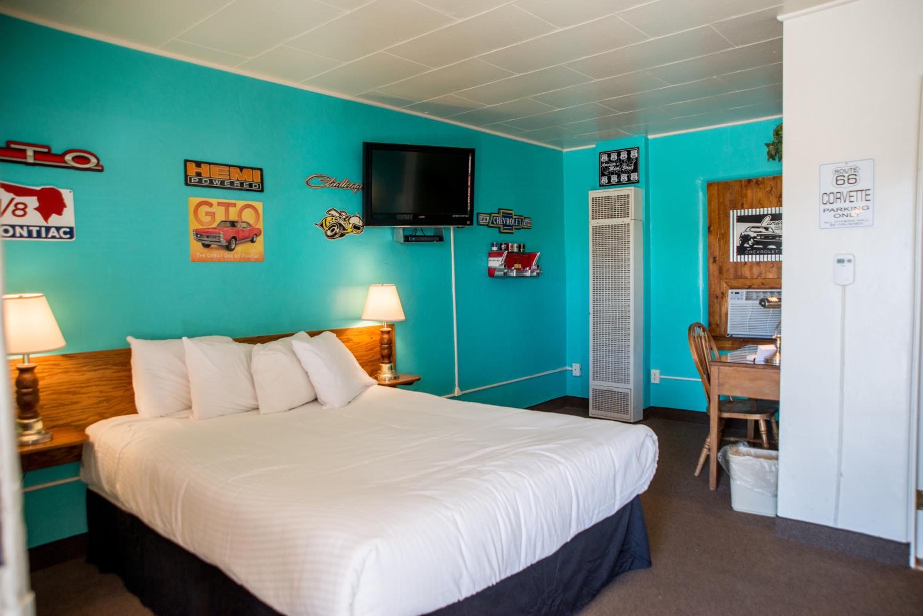A hotel room with a bed, a desk, a chair, a television, and various wall decorations including car logos and a sign indicating parking availability.