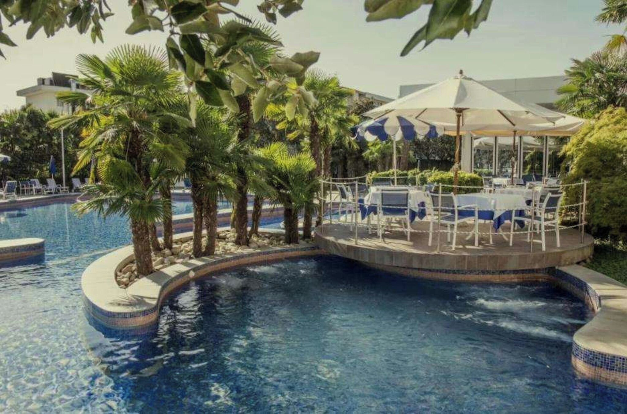 A swimming pool with a circular section, surrounded by palm trees and a covered seating area with umbrellas.
