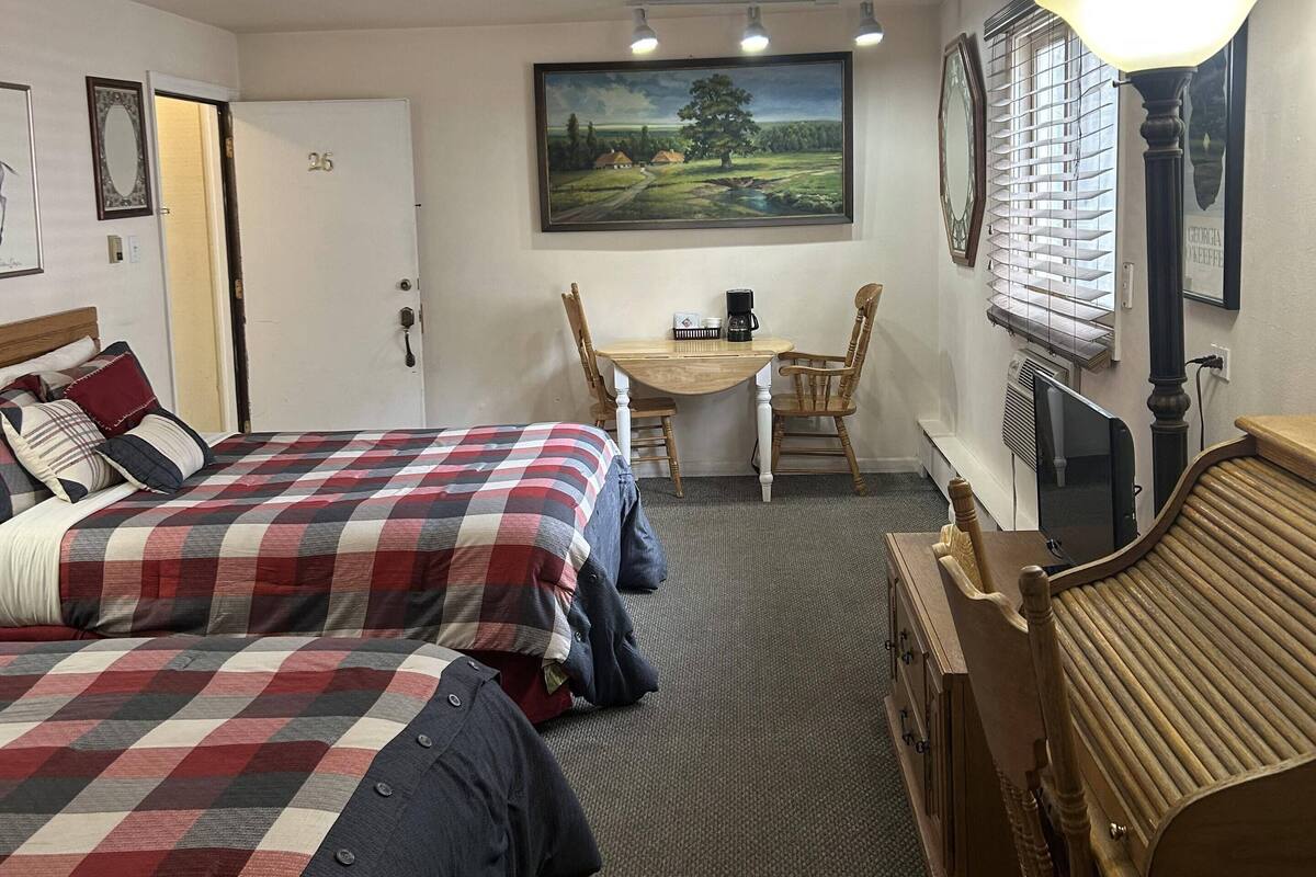 A hotel room with two beds, a desk with a chair, a lamp, a painting, and a window with blinds.