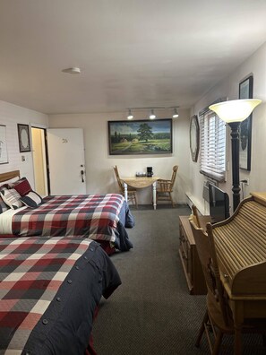 A hotel room with two beds, a desk with a chair, a lamp, a painting, and a window with blinds. - Columbine INN (Idaho Springs)