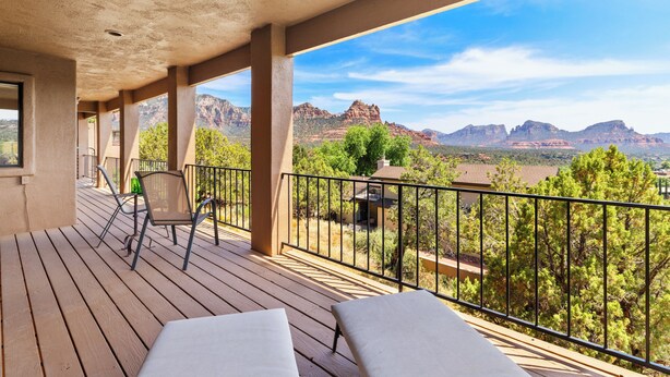 A wooden deck with two white cushioned chairs, a metal chair, and a small table. A black metal railing surrounds the deck. The deck overlooks a mountainous landscape with trees and a clear sky. - Matterhorn Inn (Sedona)