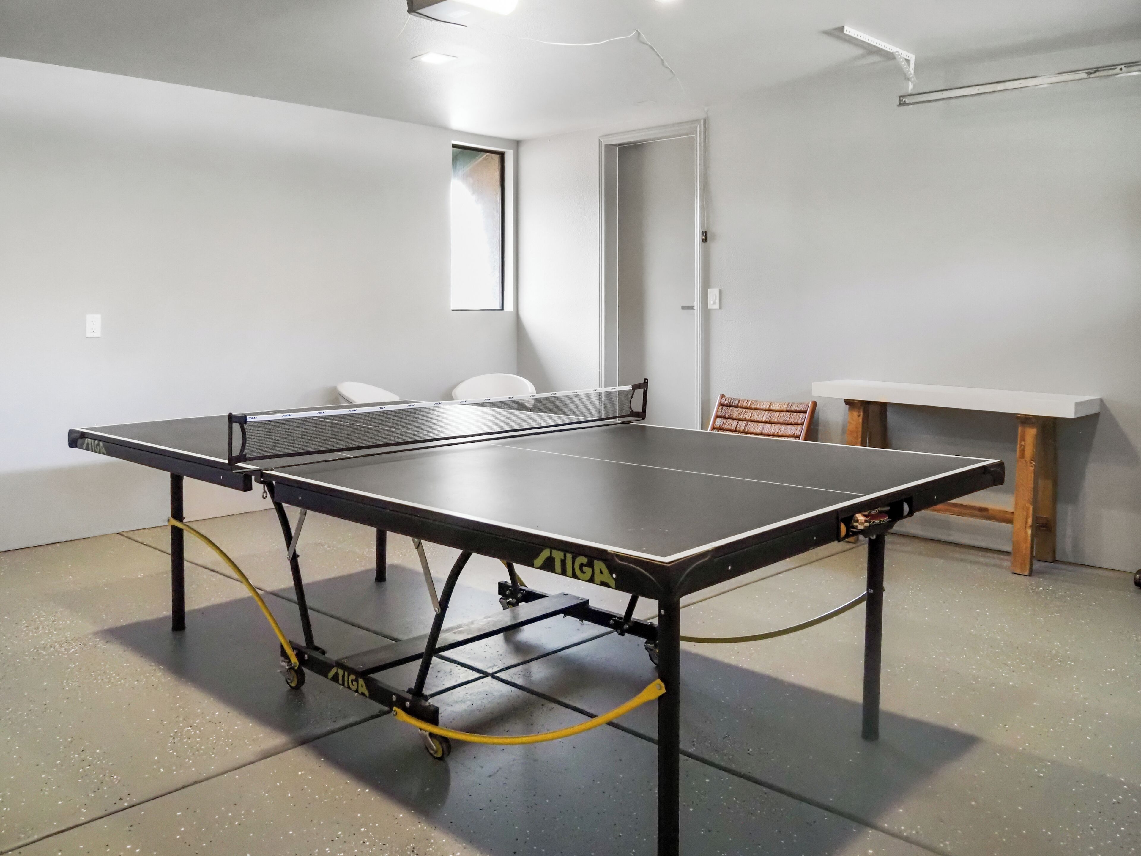 A ping pong table in a room with white walls, a door, and a small table with a chair.