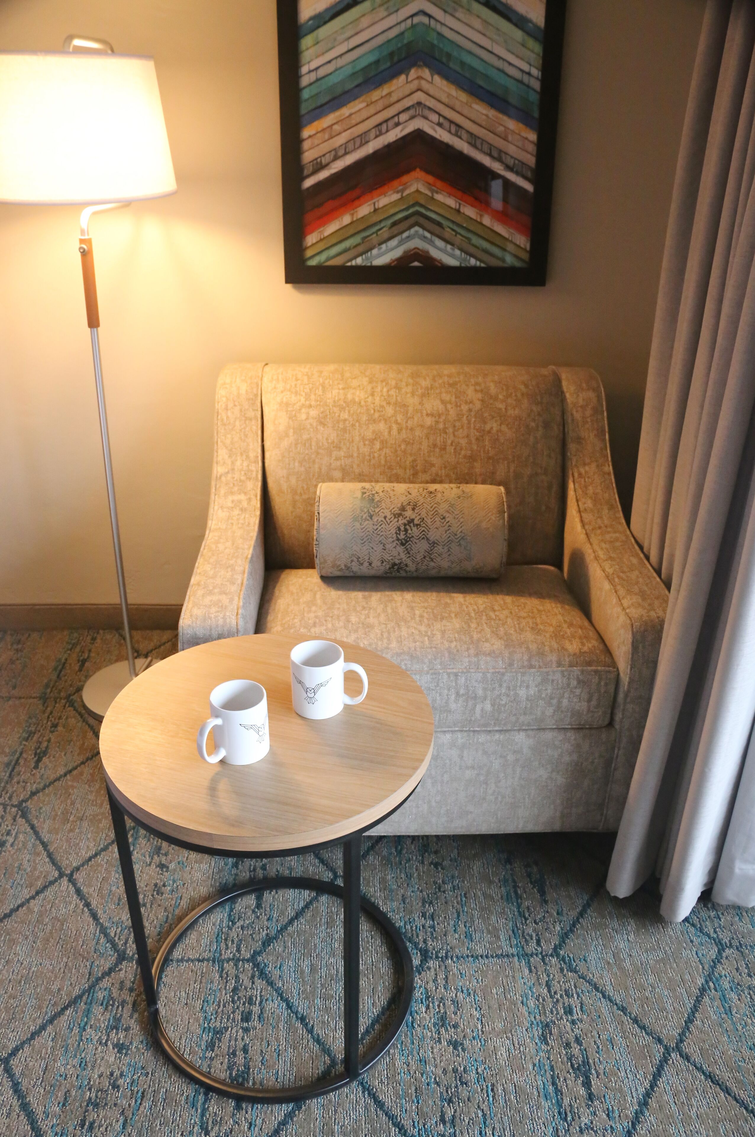 Una habitación de hotel con un sofá, una mesita redonda pequeña con dos tazas blancas, una lámpara de pie y un cuadro abstracto de colores en la pared.