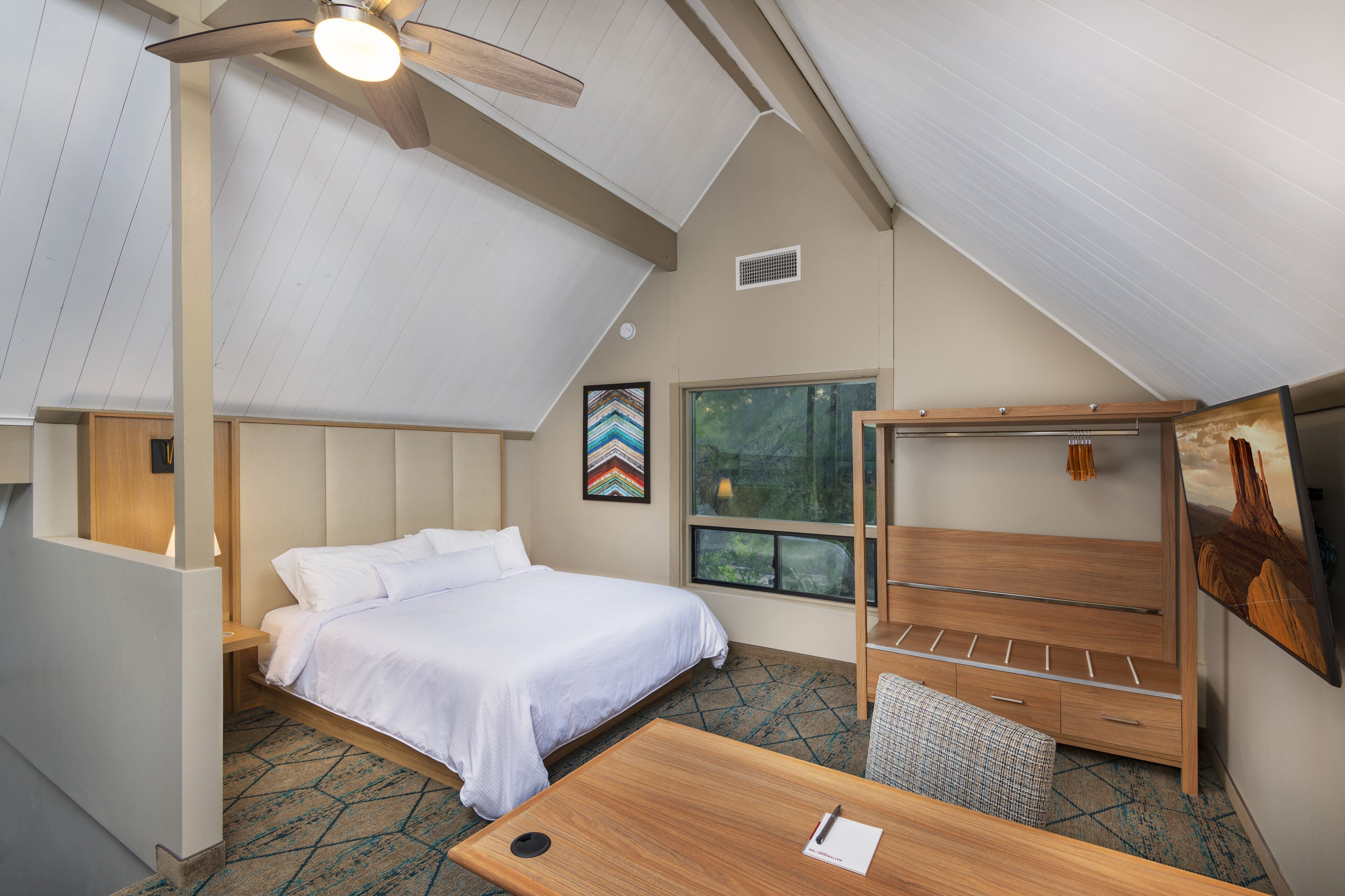 A bedroom with a bed, a desk, a chair, a ceiling fan, a window, and a painting on the wall.