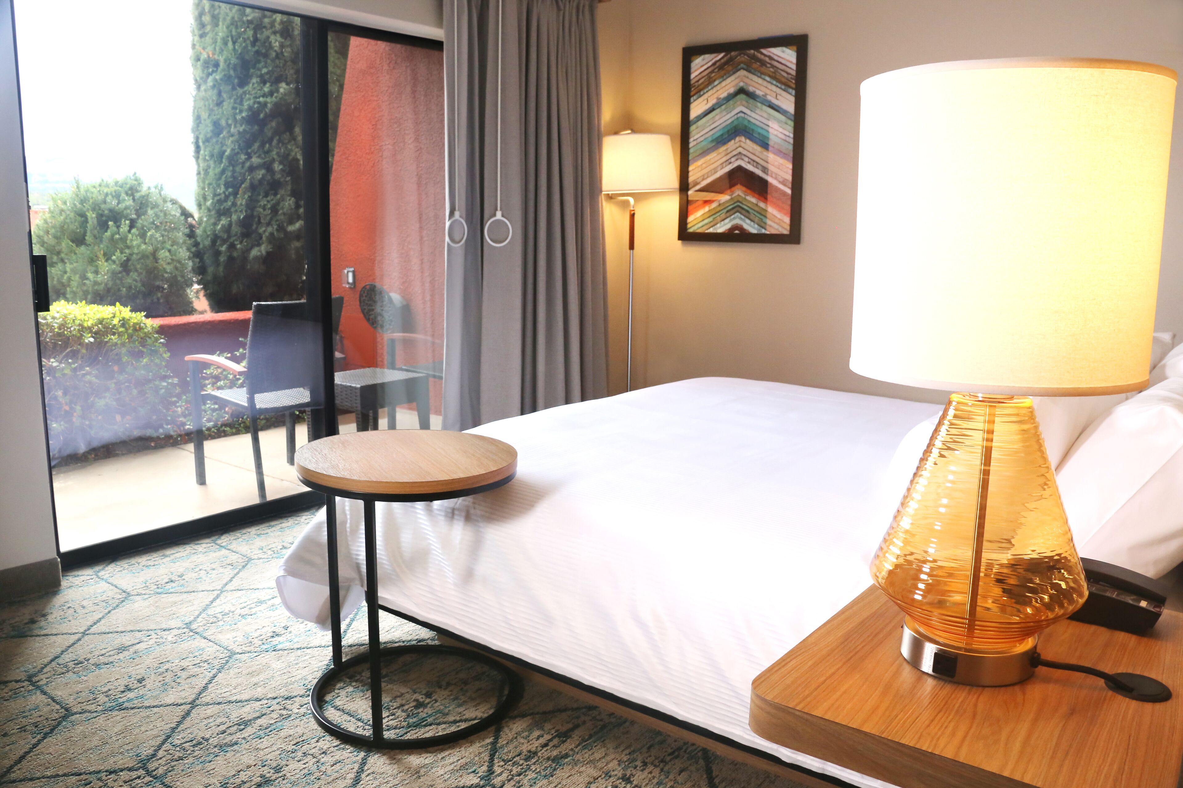 A hotel room with a bed, a round table, a lamp, a chair, and a view of the outdoors.