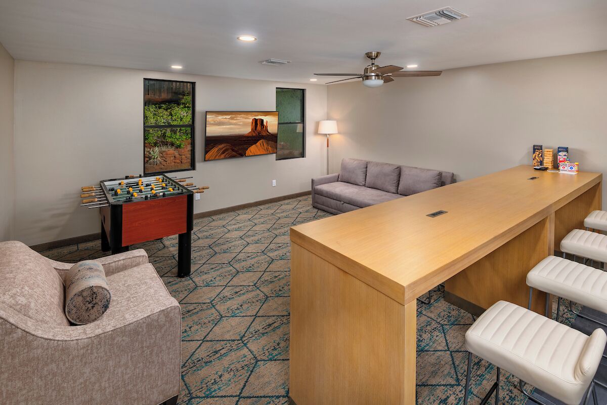 A room with a foosball table, a sofa, a bar area with stools, and a ceiling fan.