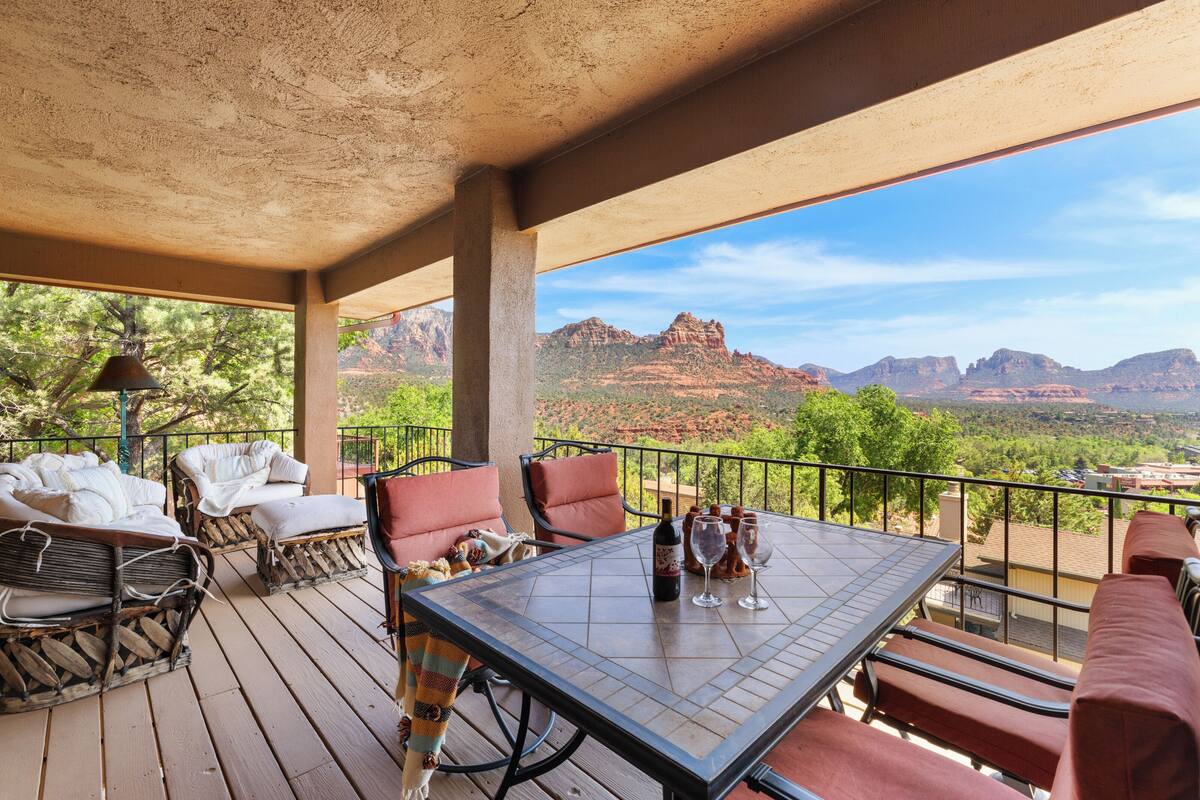 A covered outdoor seating area with wicker furniture, a table with wine glasses and bottles, and a scenic view of red rock formations.