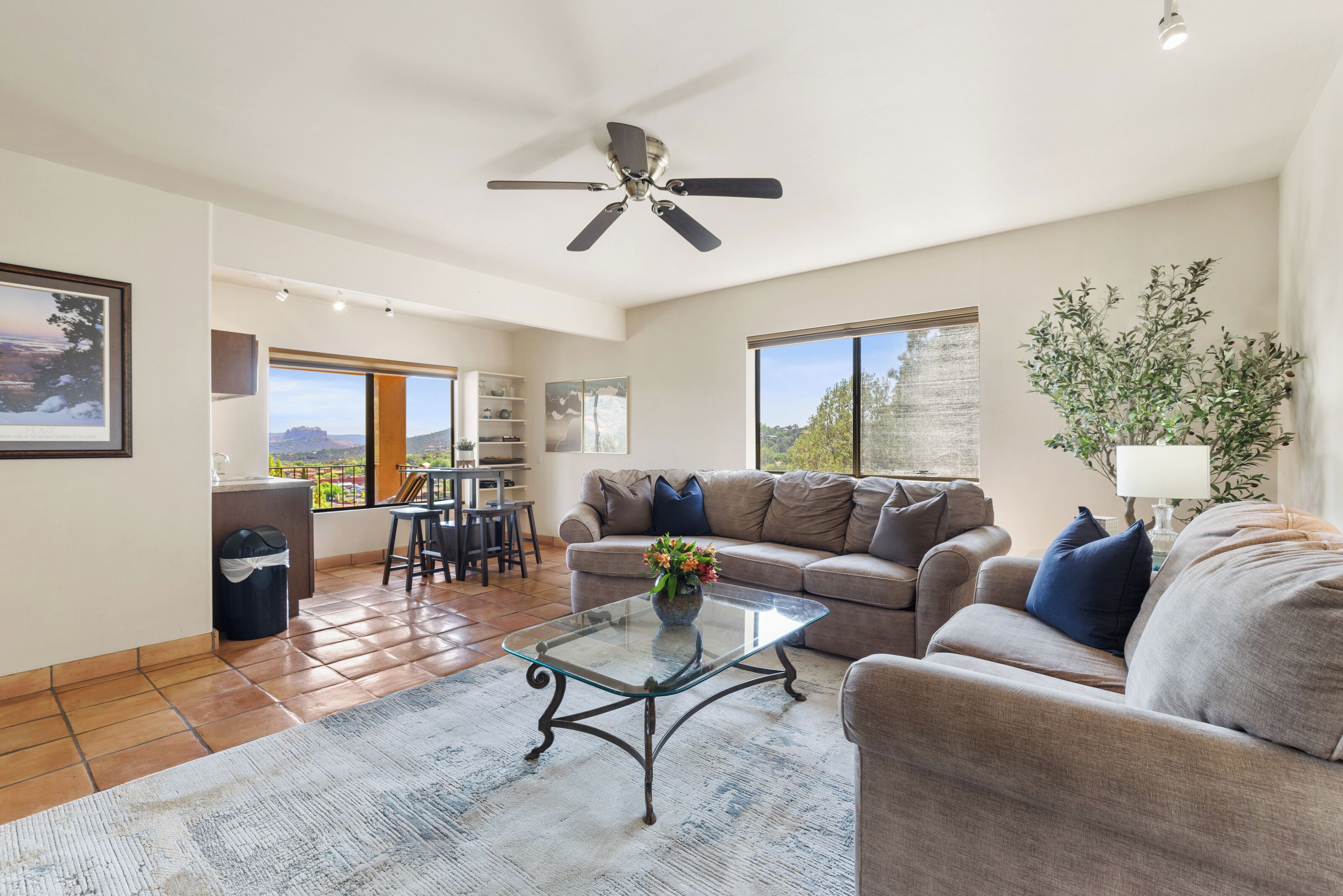 A spacious living room with a glass coffee table, a sofa, and a ceiling fan. There is a view of the outdoors through large windows.