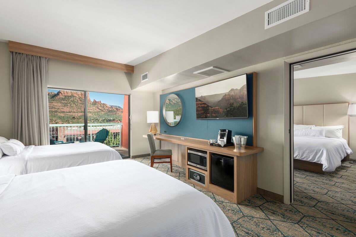 A hotel room with two beds, a desk with a coffee maker, a chair, and a large window with a mountain view.