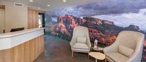 Zona de recepción con un mostrador de madera, dos sillones y una pequeña mesa. Se observa un gran mural con un paisaje montañoso al fondo.