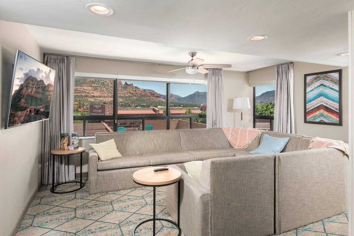 A modern hotel room with a large window offering a view of mountains and a cityscape. There is a gray sofa, a small round table, and a ceiling fan.