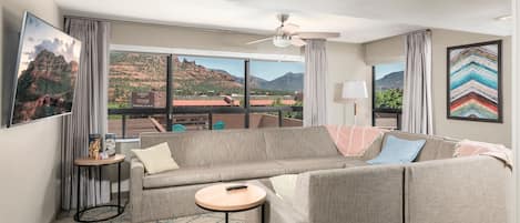 A modern hotel room with a large window offering a view of mountains and a cityscape. There is a gray sofa, a small round table, and a ceiling fan.