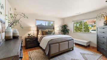 A bedroom with a bed, bedside tables, a bench, a dresser, and a window with a view of trees and a car.