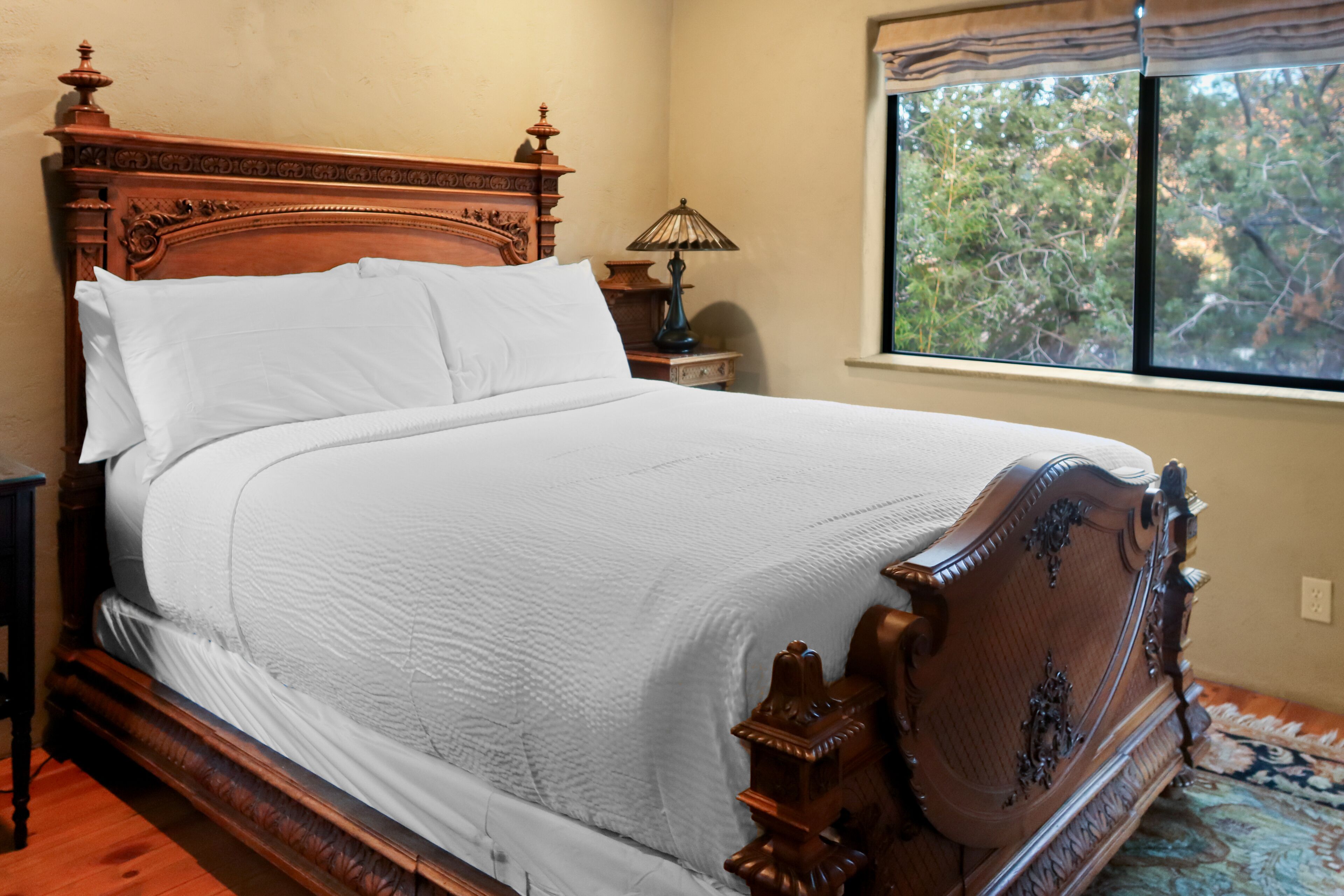 A neatly made bed with a wooden headboard and footboard, a bedside table with a lamp, and a window with a view of trees.