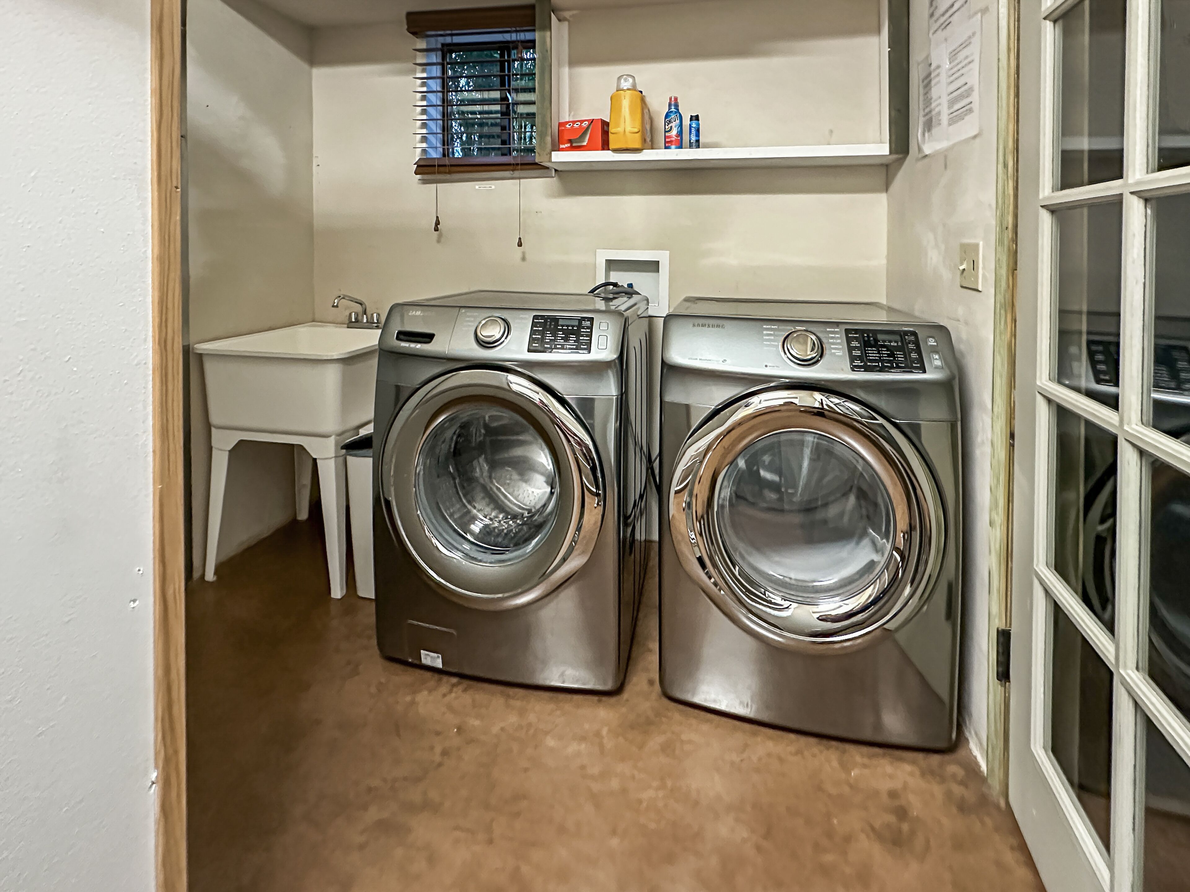 A laundry room with two front-loading washing machines, a small sink, and a window with blinds.