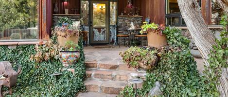 A stone staircase leading to a wooden door with a glass panel, surrounded by lush greenery and potted plants.