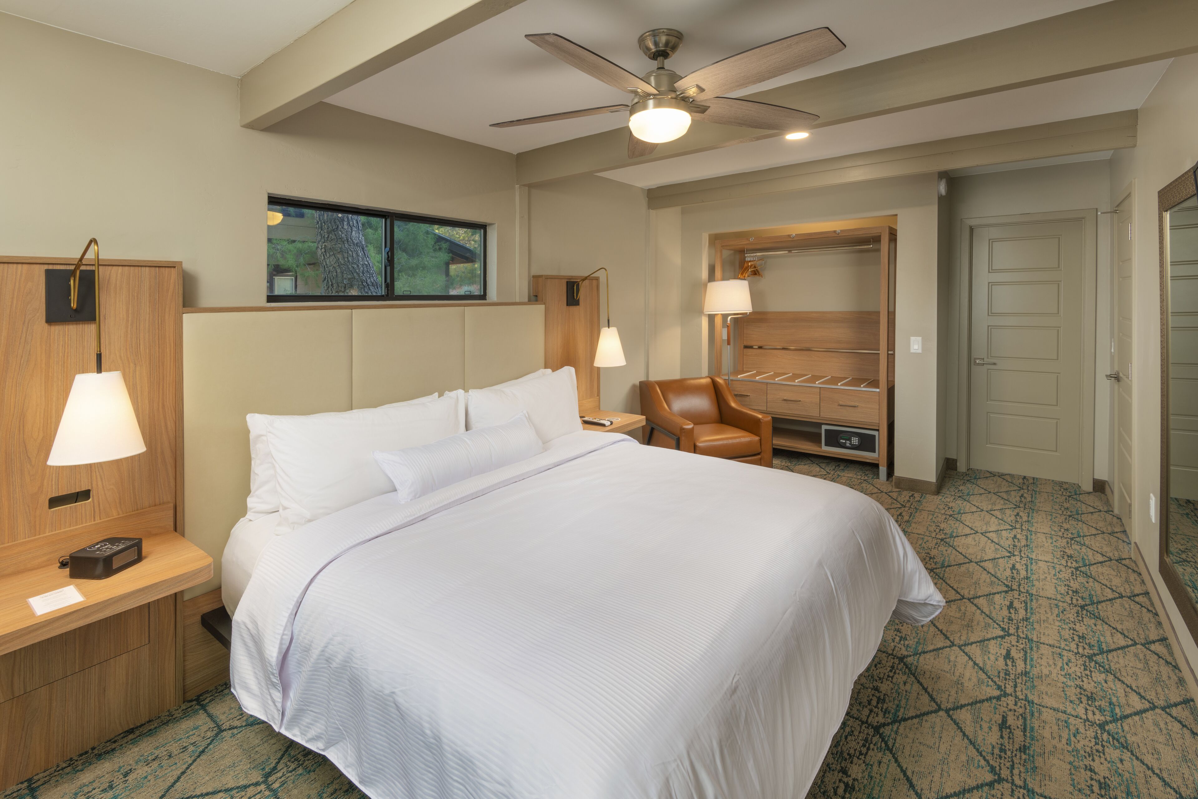 Ein Hotelzimmer mit einem großen Bett, einem Deckenventilator, einem Holzschrank, einem Sessel und einem Fenster mit Blick auf Bäume.